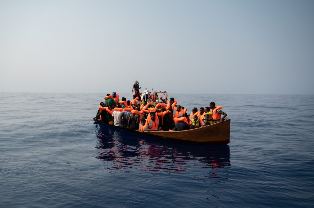 Es ist ein seeuntüchtiges Boot abgebildet, das überfüllt ist. Die Menschen haben Schwimmwesten an und werden von der Crew auf einem Rettungsboot angeleitet.